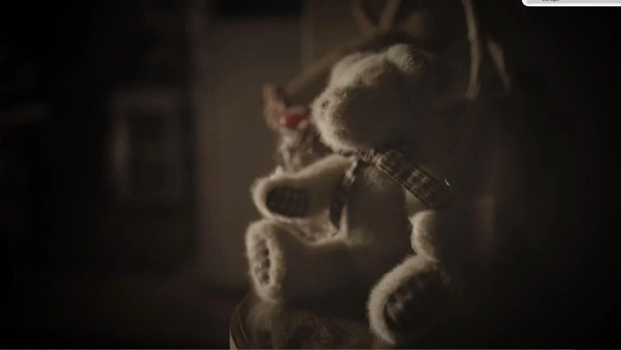Murder at Carter County Mines (2023) movie still: closeup — A plush teddy bear with a plaid ribbon sits on a small round table. The background is…