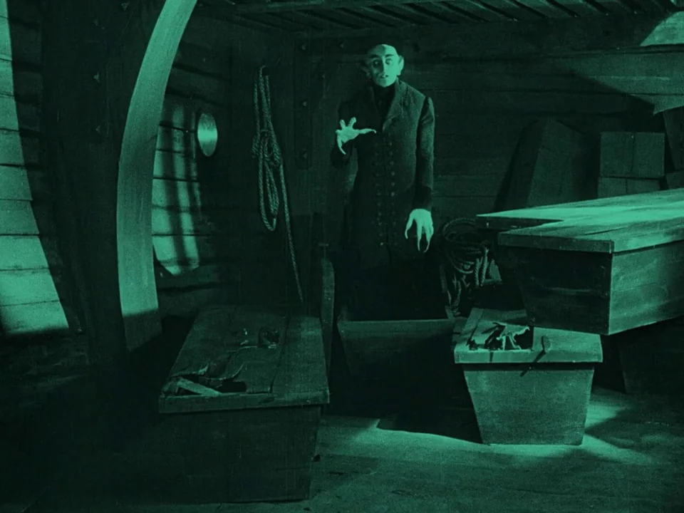 Nosferatu (1922) movie still: medium — A dark wooden ship hold is cluttered with several large rectangular wooden crates. A tall, pale man in a long…