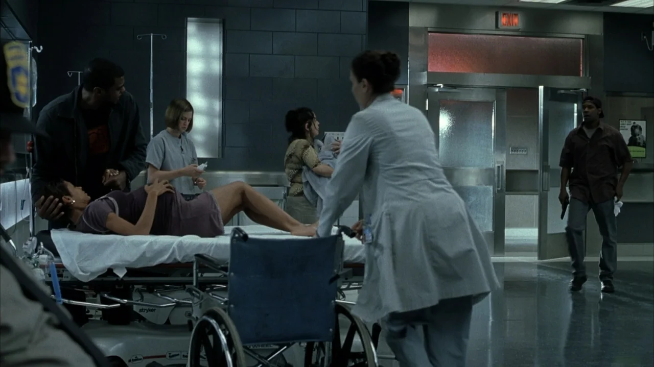 John Q (2002) movie still: wide — A sterile hospital emergency room features dark tiled walls and glass double doors. Several people congregate…