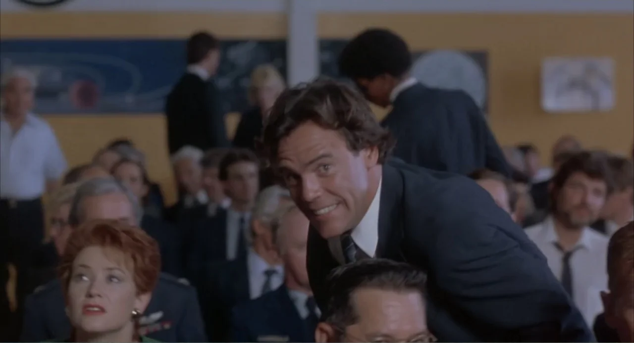 Making Mr. Right (1987) movie still: medium — A crowded auditorium features yellow walls and rows of people in professional attire. A man leans over…