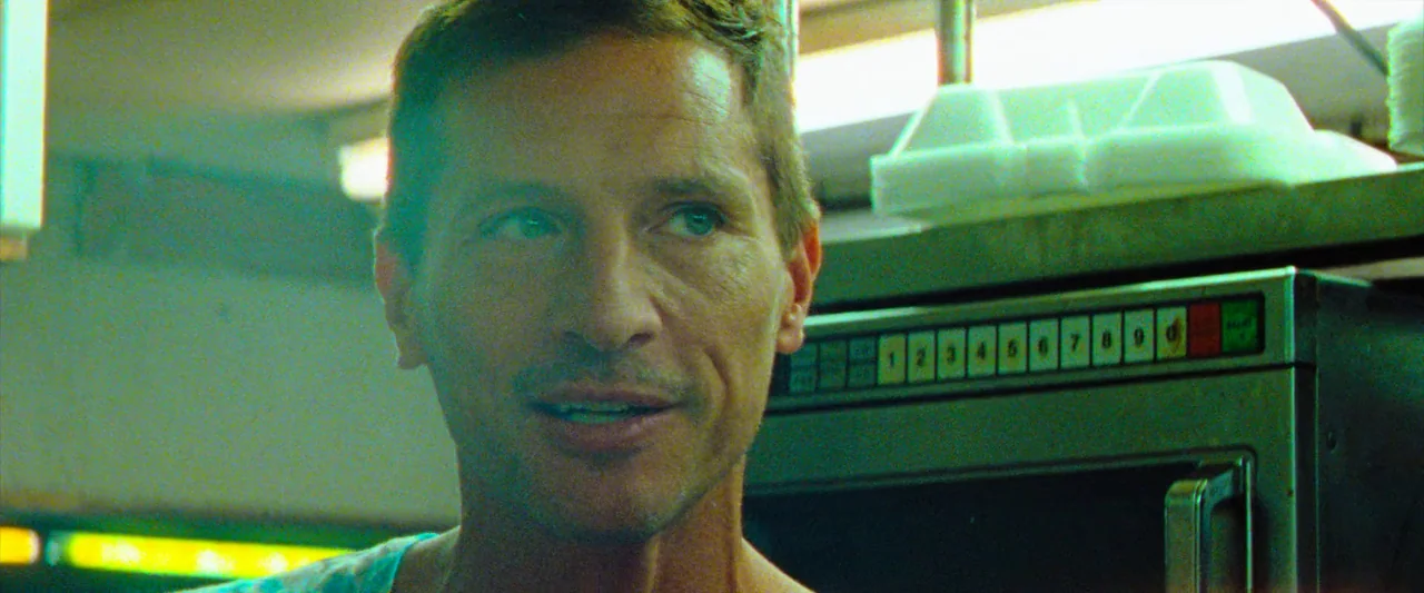 Red Rocket (2021) movie still: closeup — A man stands before a commercial microwave in a convenience store. The setting includes a stack of white…