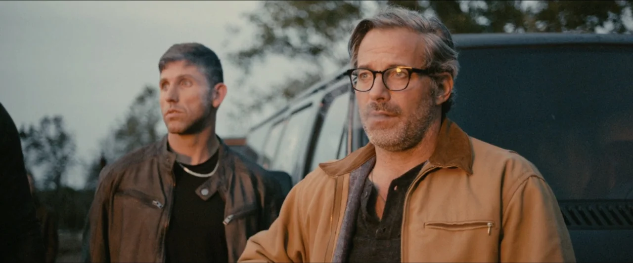Ambush (2020) movie still: medium — Two men stand outdoors in an open space during sunset. They look toward the side while standing near a dark…