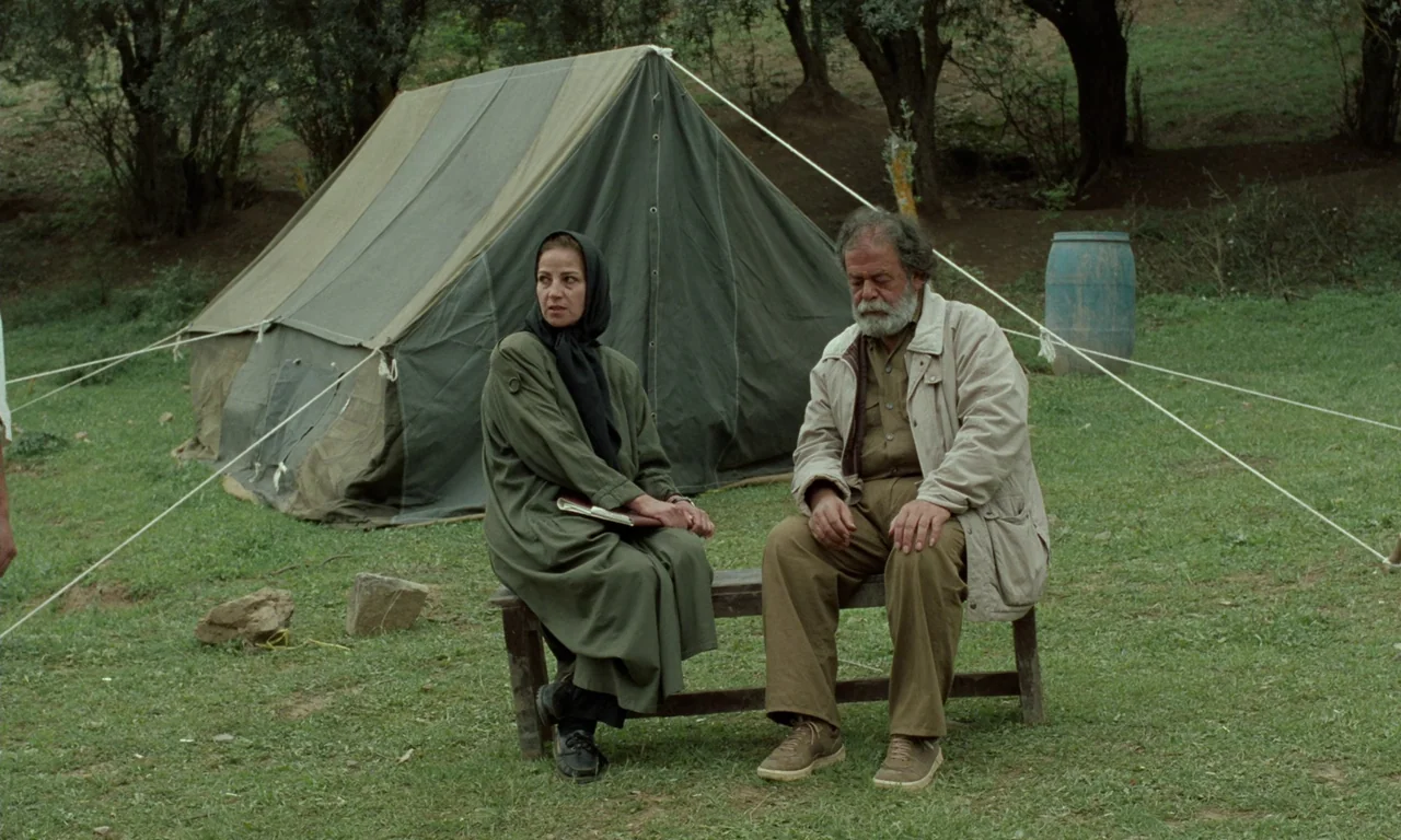 Through The Olive Trees (1994) movie still: fullbody — A grassy clearing features a military-style canvas tent pitched among scattered trees. A man…