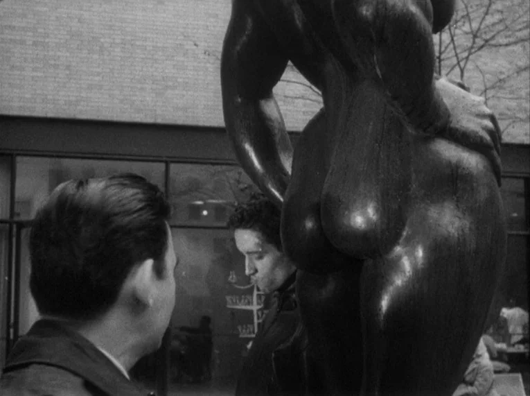 Shadows (1958) movie still: medium — An outdoor paved area features a large, dark, abstract bronze statue of a figure. Two men stand close to the…
