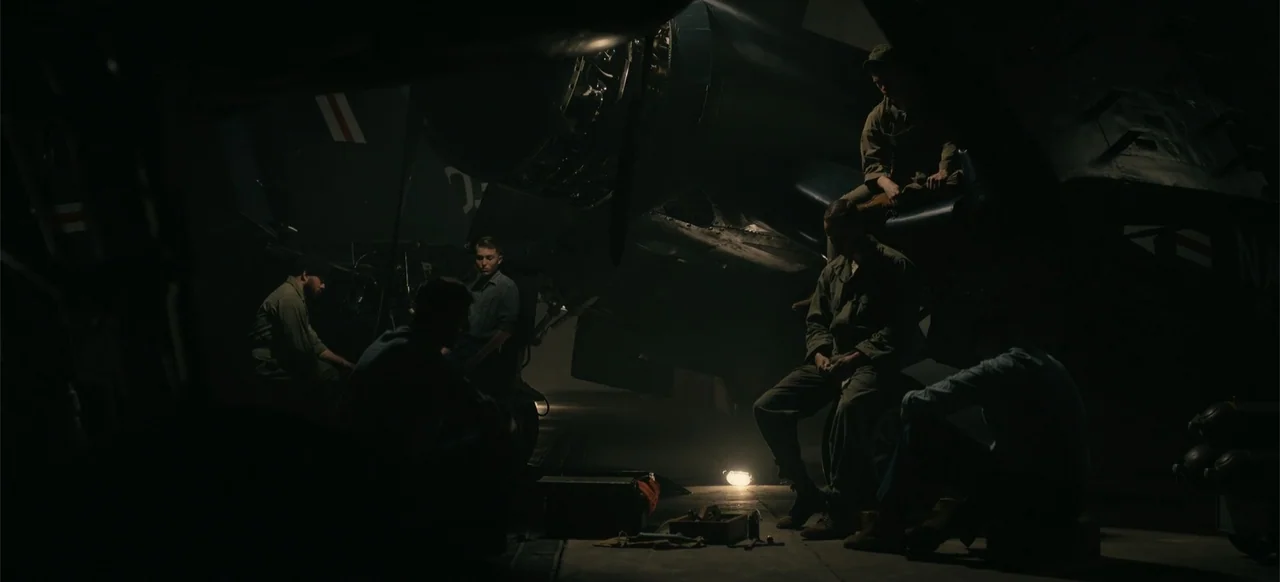 Devotion (2022) movie still: wide — A dark military aircraft hangar contains several men sitting near a large parked plane. The men rest in the…