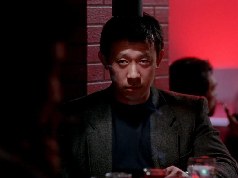 Black Snow (1990) movie still: medium — A dimly lit indoor space features a red-painted brick wall background. A man sits at a table and stares…