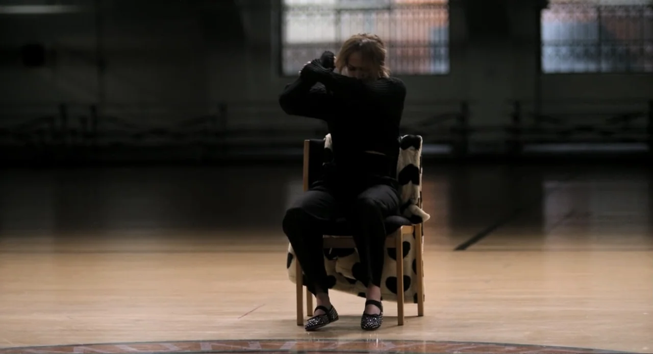 The Greatest Love Story Never Told (2024) movie still: medium — A spacious wooden basketball court interior features a single chair draped with a…