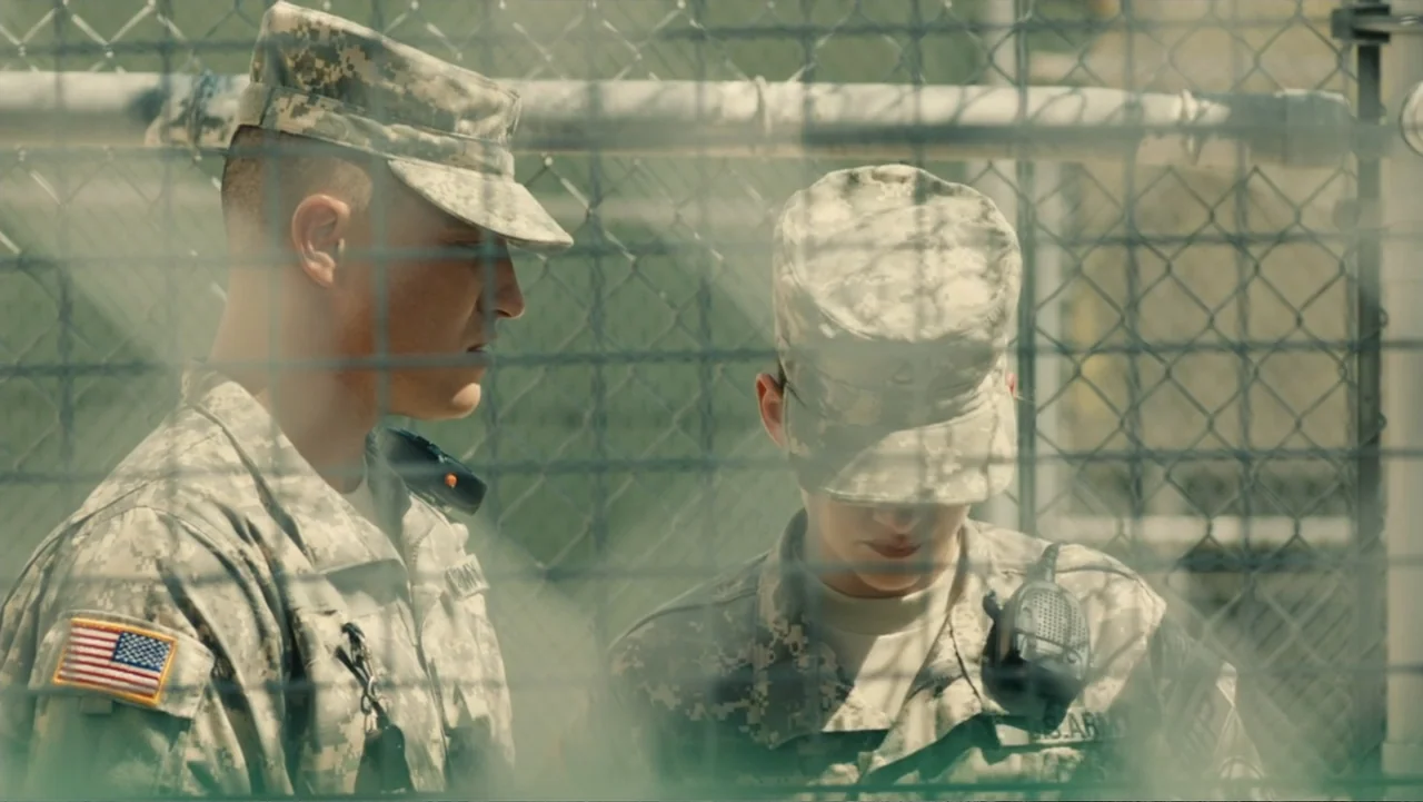 Camp X-Ray (2014) movie still: medium — Two US Army soldiers in combat fatigues stand behind a metal chain link fence. They look downward while…