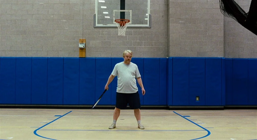The Savages (2007) movie still: fullbody — An indoor gymnasium features light-colored flooring and walls covered in blue protective padding. A…