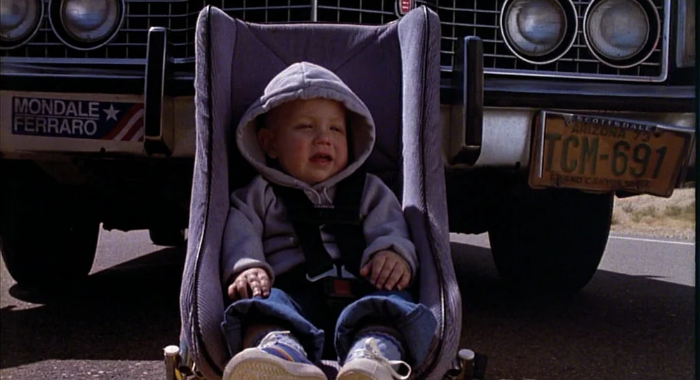 Raising Arizona (1987) movie still: medium — A toddler sits in a car seat placed on the asphalt beside a car. The vehicle bumper has a Mondale…