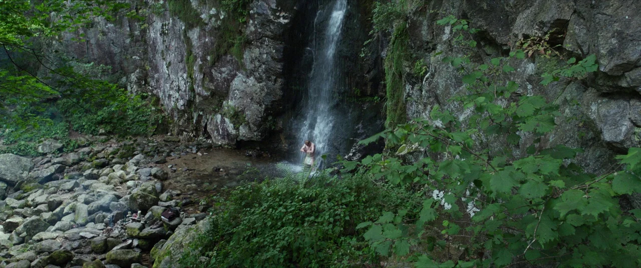 The Wailing (2016) movie still: wide — A secluded rocky gorge features a tall thin waterfall cascading over dark craggy stone. A solitary man stands…