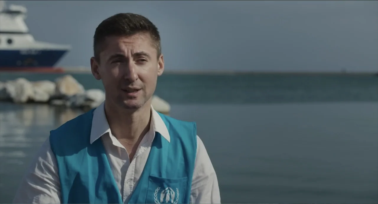 Human Flow (2017) movie still: medium — A man stands outdoors near a waterfront with a ship visible in the distance. He wears a blue utility vest…