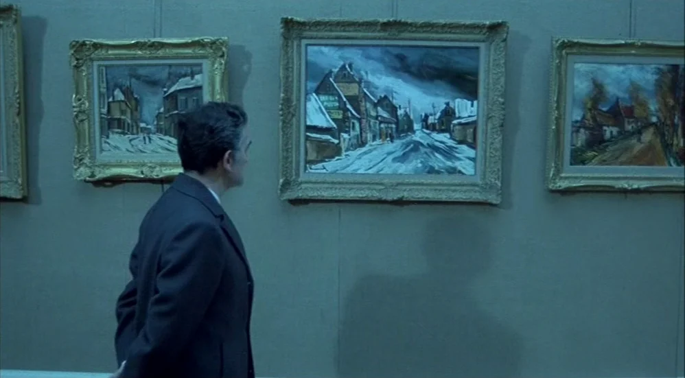 Un Flic (1972) movie still: medium — An art gallery wall features several framed paintings depicting snowy landscapes hung in a row. A man stands in…