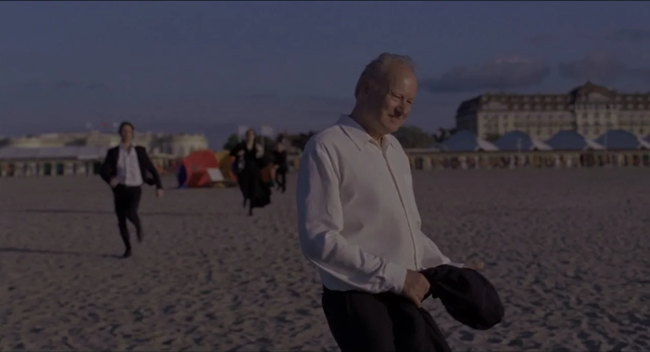 Sentimental Value (2025) movie still: medium — A wide sandy beach stretches out under a clear sky during the golden hour. An older man in a white…