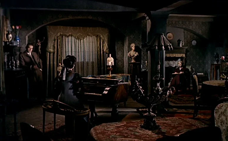 Mill of the Stone Women (1960) movie still: wide — A dimly lit room features ornate furniture, heavy curtains, and patterned wallpaper from the…