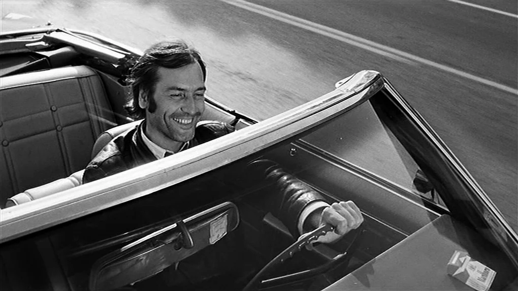 The State of Things (1982) movie still: medium — A man drives a vintage convertible on an asphalt road during the day. The man sits in the driver's…