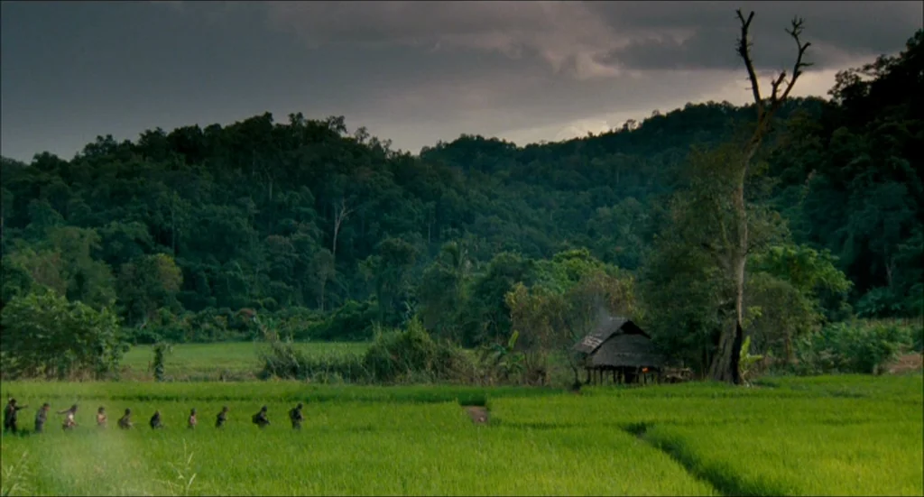 Rescue Dawn movie still: wide — A wide view of a vibrant green rice paddy beneath a dark, cloudy sky. A group of soldiers traverses the field in a…