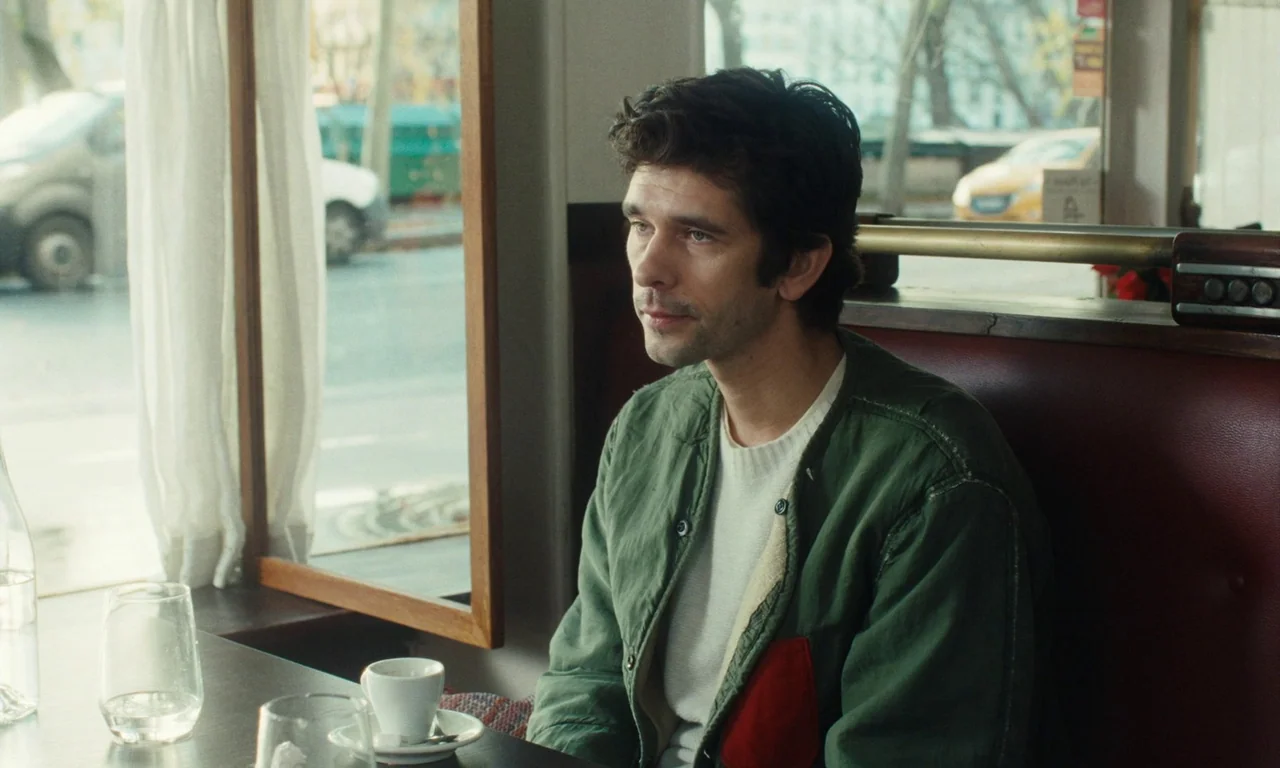 Passages (2023) movie still: medium — A man sits at a table inside a diner booth near a large glass window. Outside, a blurred urban street scene is…