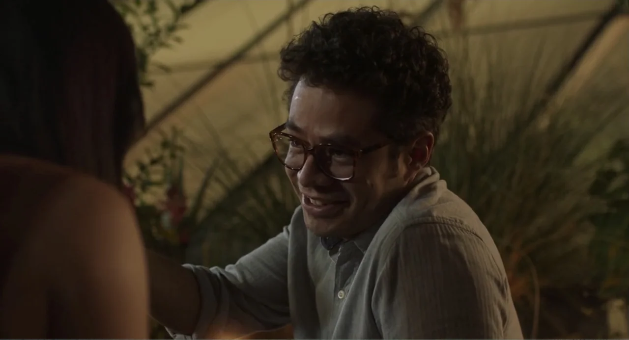 Casando a mi Ex (2023) movie still: medium — A man sits inside a greenhouse filled with tropical foliage at night. He smiles while speaking to a…