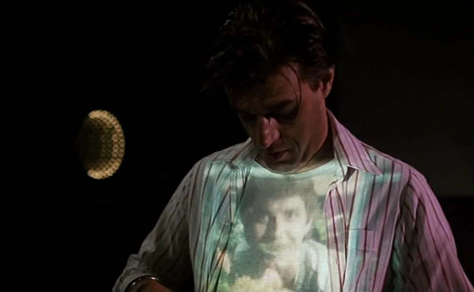 The Fourth Man (1983) movie still: medium — A man stands in a dark space, his chest illuminated by a bright projection. He gazes downward at the…