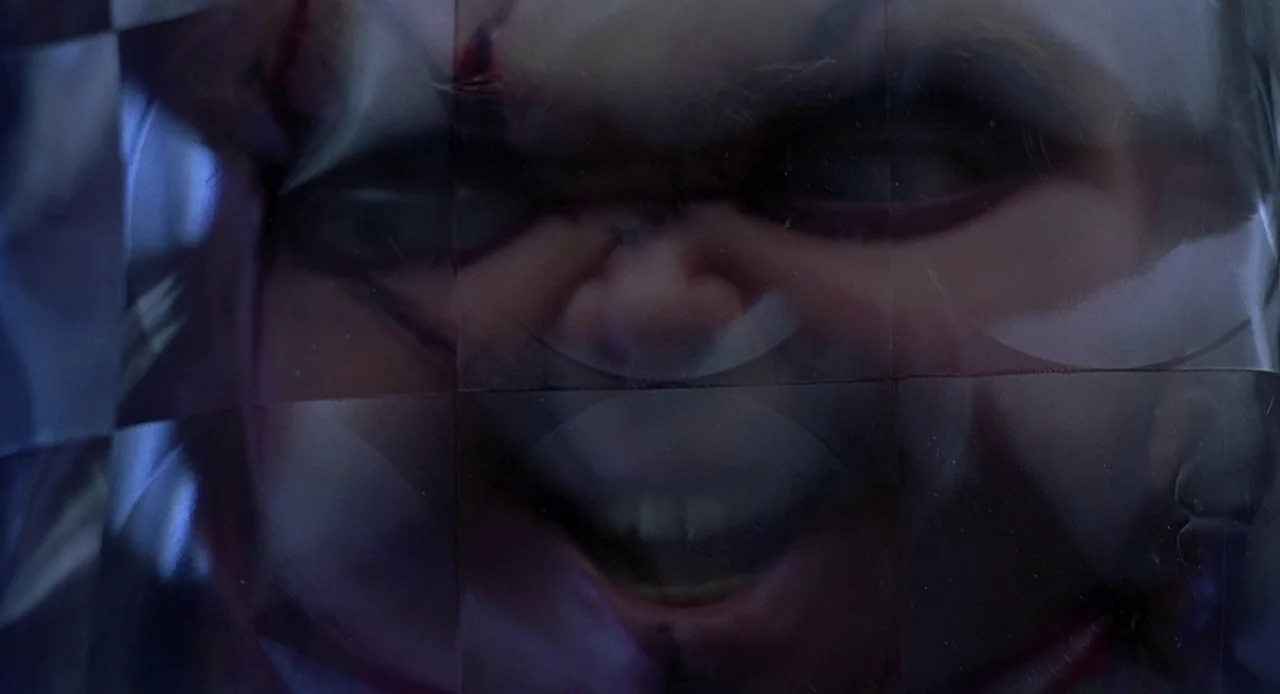 Bride of Chucky (1998) movie still: closeup — A doll's face is distorted by a layer of textured clear plastic. The image is seen through the warped…