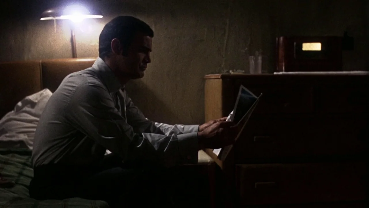 Bullitt (1968) movie still: medium — A dimly lit room contains a bed, a wooden dresser, and a desk lamp. A man sits on the side of the bed while…