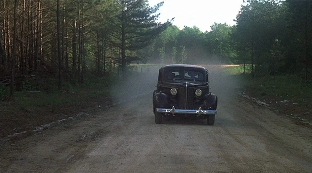 Thieves Like Us (1974) movie still: wide — A dark vintage automobile drives down a narrow dirt road surrounded by dense woods. The car kicks up a…