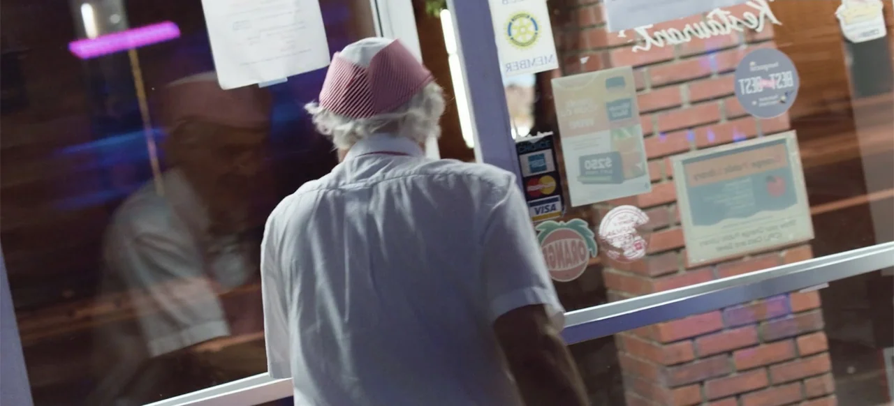 Hellbreeder (2004) movie still: medium — A glass door with various decals and signage covers the view of a retail space. An elderly man in a white…