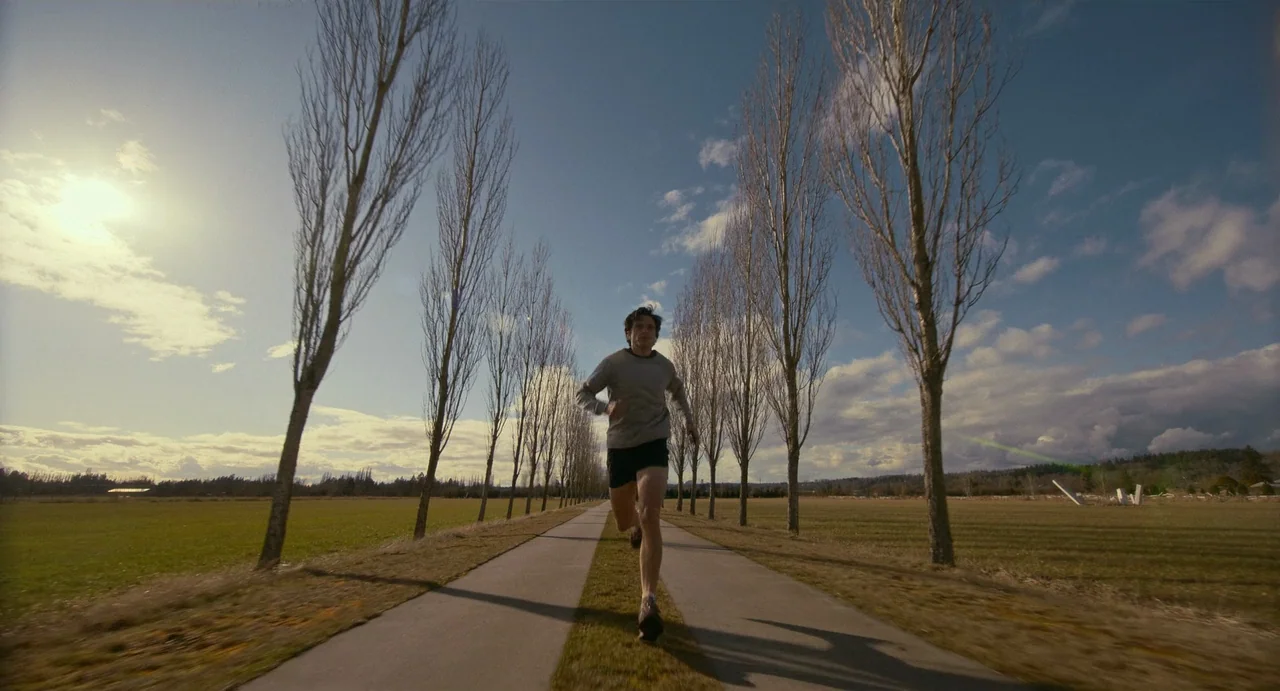 Fresh (2022) movie still: fullbody — A long paved path stretches through a rural field lined with tall bare trees. A man in athletic gear jogs…