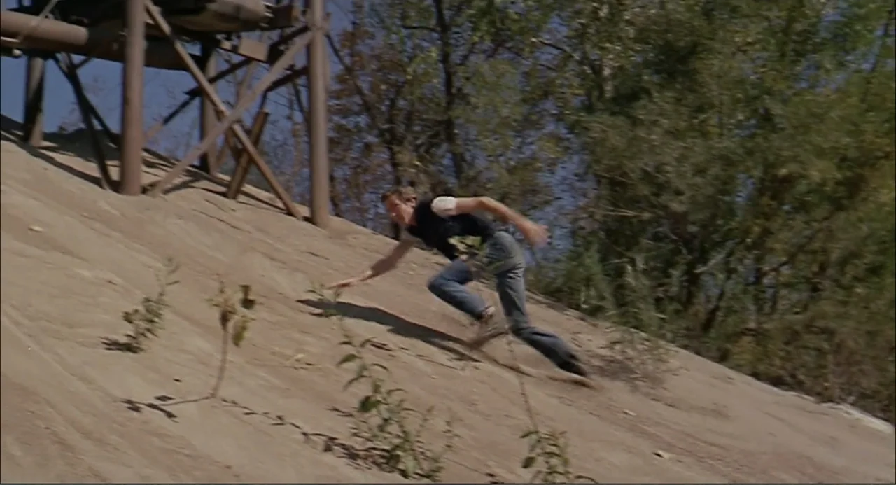 In the Heat of the Night (1967) movie still: wide — A steep, sandy industrial embankment borders an area with sparse vegetation and trees. A young…