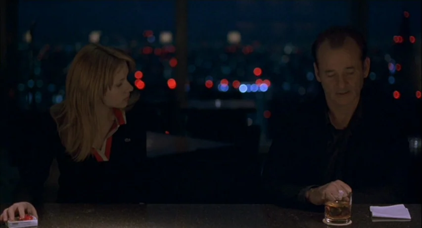 Lost In Translation (2003) movie still: medium — A dark bar interior overlooks a blurred city skyline with distant red and blue lights. A man and…