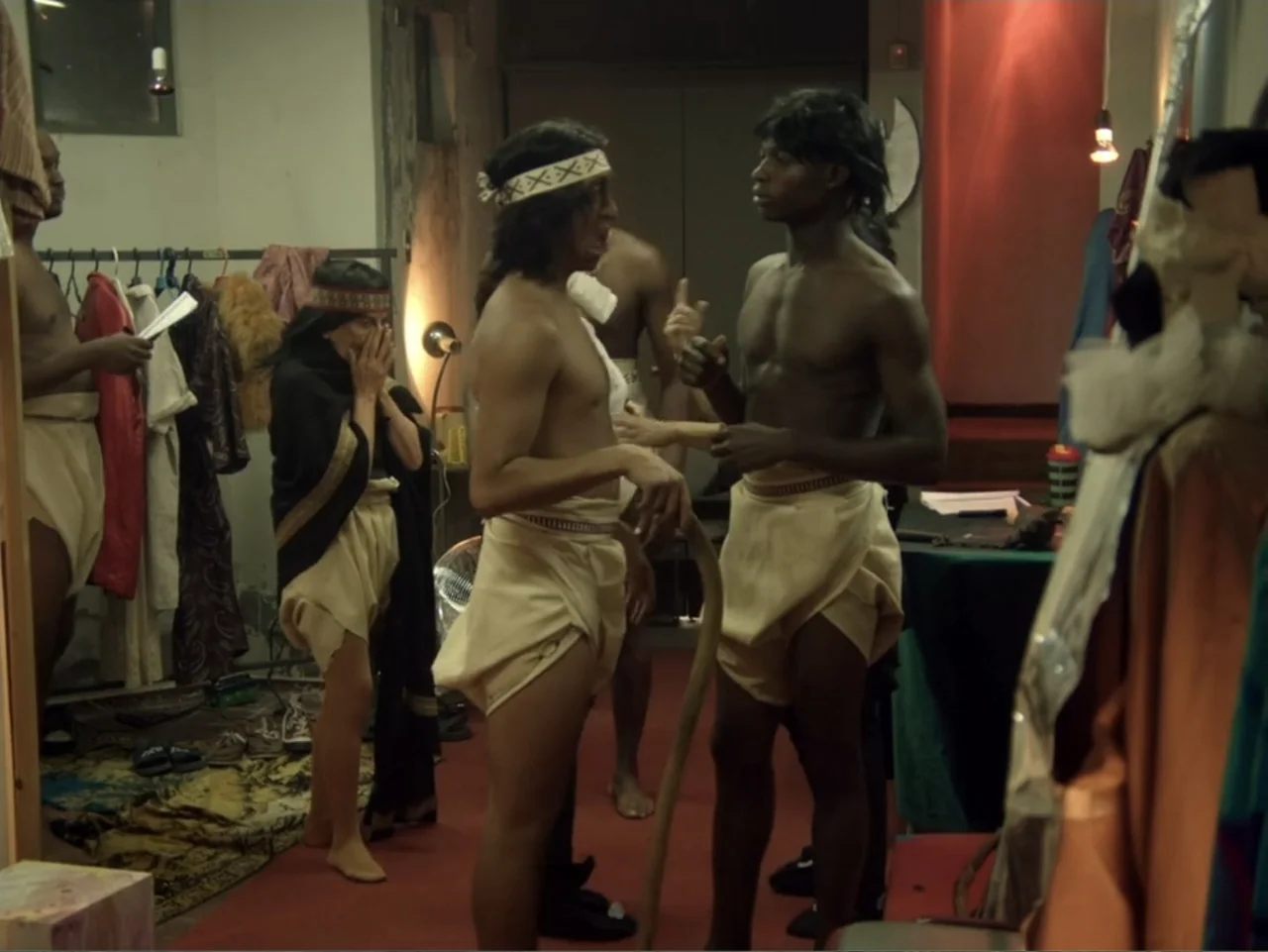 History and Geography (2024) movie still: medium — A cluttered backstage dressing room filled with costume racks, hanging fabrics, and scattered…