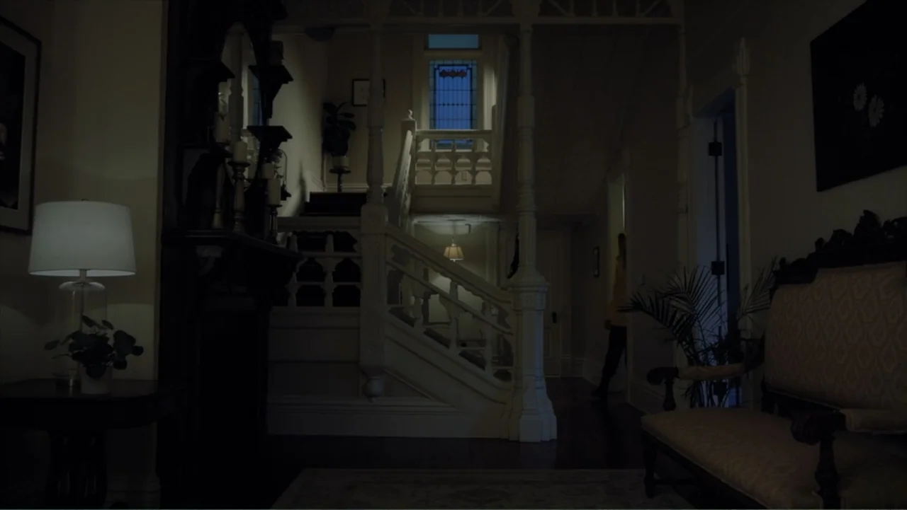Storm Damage (2000) movie still: wide — A dimly lit Victorian-style foyer with a grand white staircase and ornate woodwork. A single figure walks…