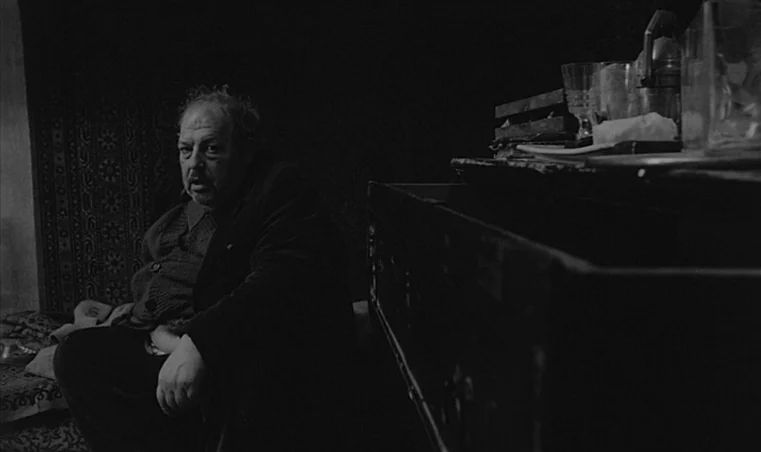 Sátántangó (1994) movie still: medium — A dark, sparsely lit room contains a person sitting beside a wooden chest. The chest top is cluttered with…