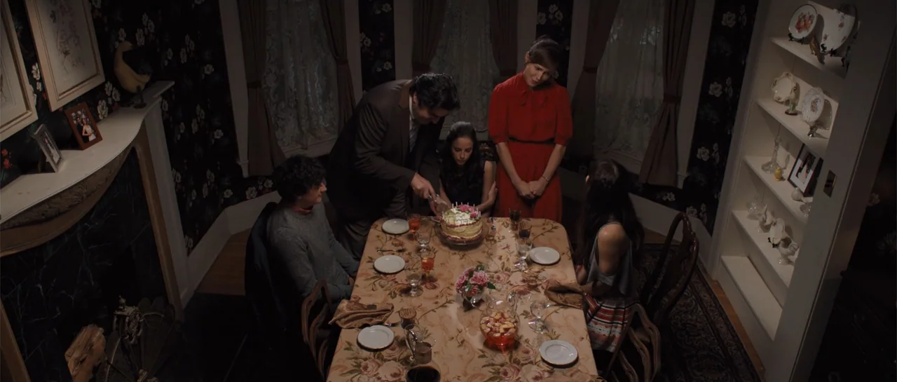 The Truth About Emanuel (2013) movie still: wide — A formal dining room features floral wallpaper and a table set for a meal. A man in a suit cuts a…