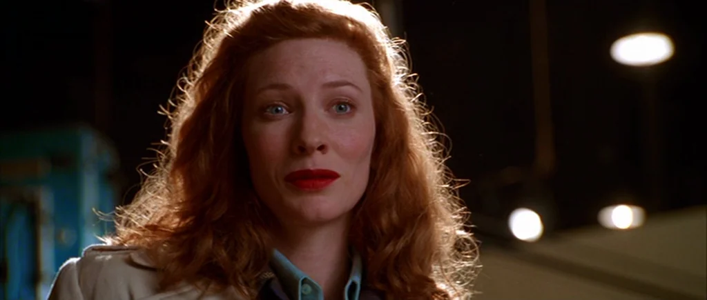 The Aviator (2004) movie still: closeup — An interior space with industrial lighting sources visible in the dark background. A woman with wavy red…