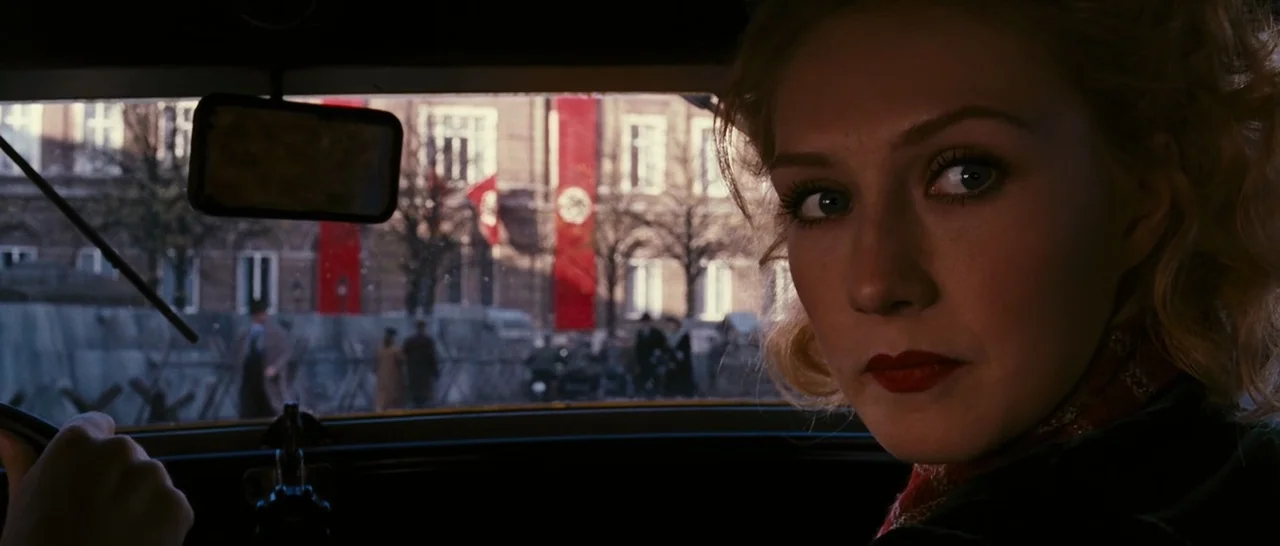 Black Book (2006) movie still: closeup — The interior of a vintage car provides a dark frame for an exterior city street. Nazi banners hang on the…