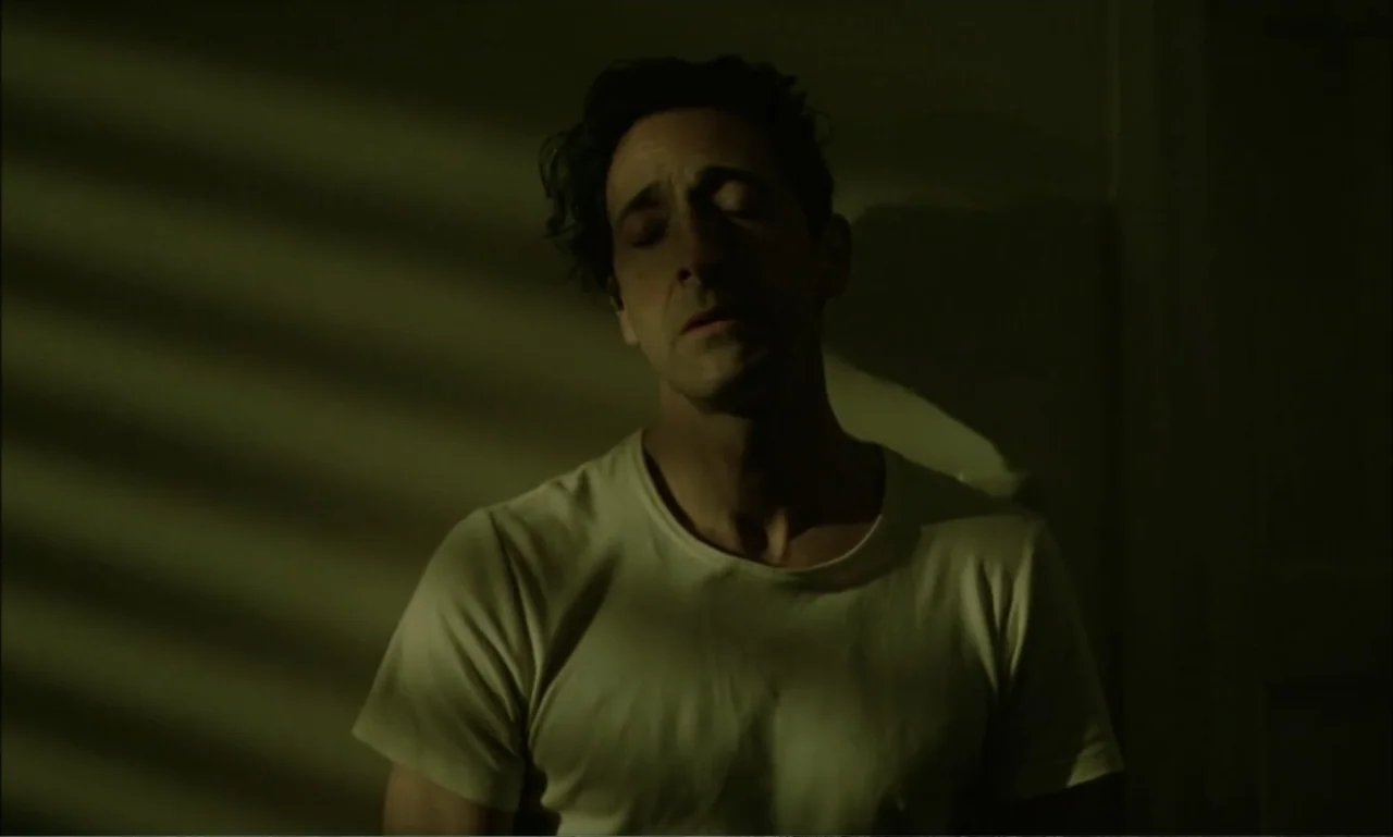 The Brutalist (2024) movie still: medium — A dark indoor space is lit by light filtering through slatted window blinds. A man in a white t-shirt…