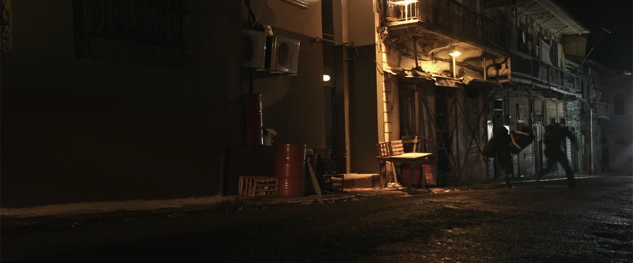 Kimura (2017) movie still: wide — An urban residential street at night features multi-story wooden buildings with overhanging balconies. Two figures…