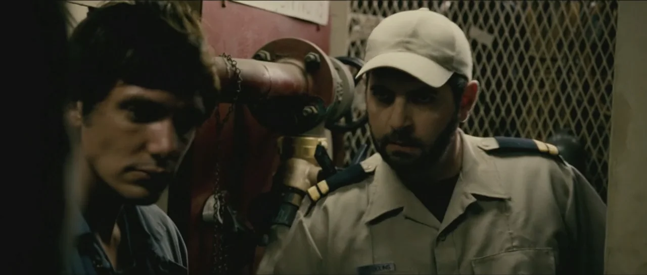 Contraband (2012) movie still: medium — A utilitarian space featuring industrial piping and a metal mesh wall in the background. Two men are…