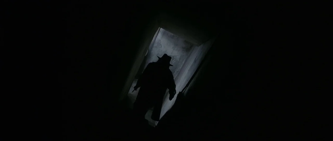 Bones movie still: wide — A dark, narrow stone staircase descends into a dimly lit basement area. A solitary man in a fedora and trench coat walks…