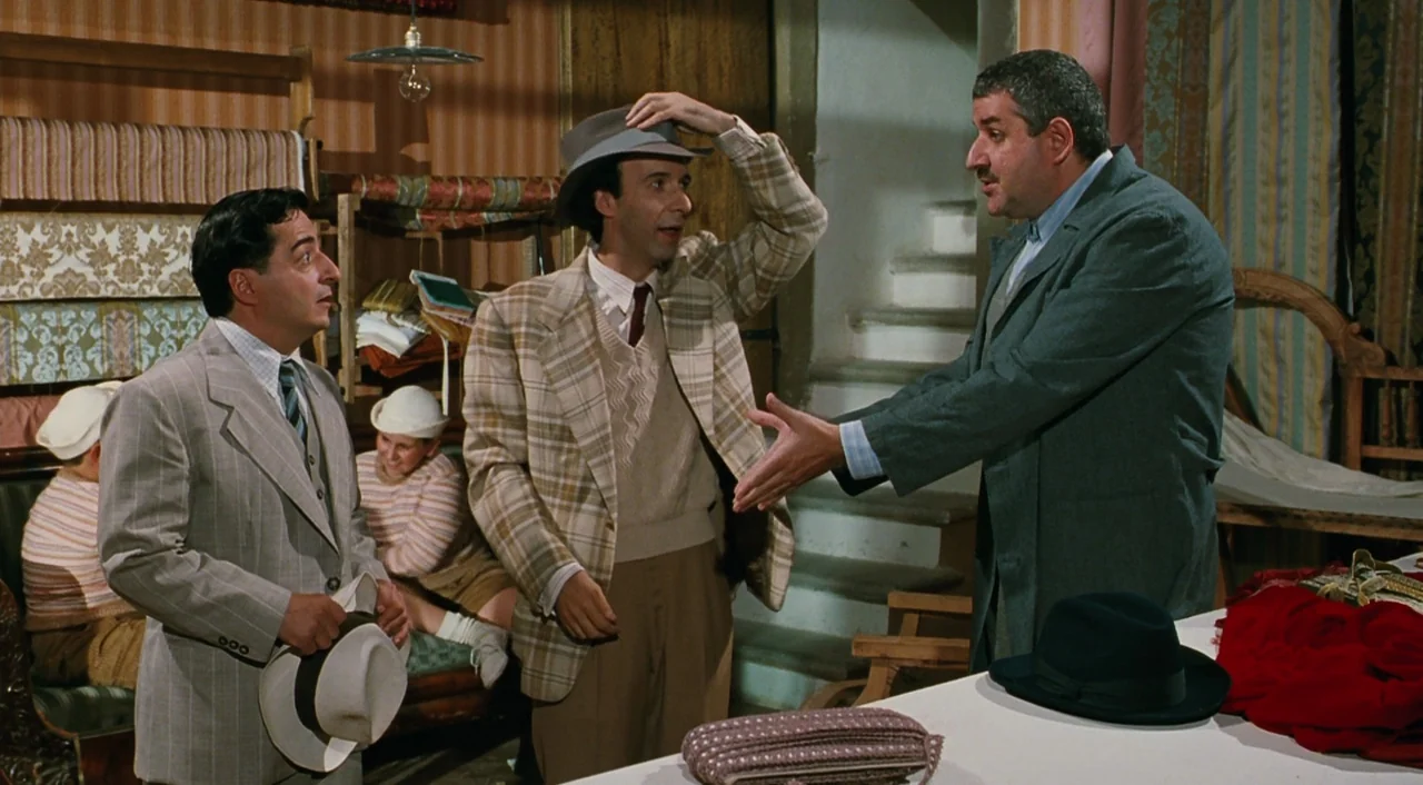 Life is Beautiful (1997) movie still: medium — A cluttered shop interior features rolls of decorative fabric stacked against back walls. Three men…