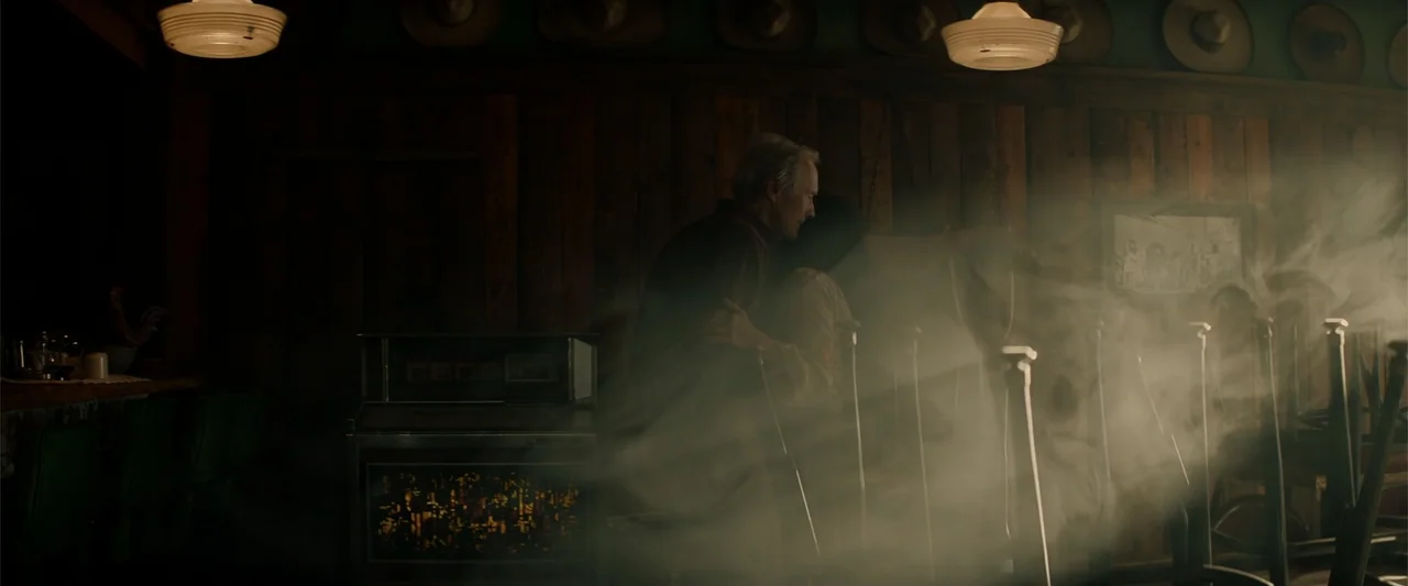 Cry Macho (2021) movie still: medium — A rustic bar interior features wood paneling and hats hung on the wall. An elderly man and a woman embrace…