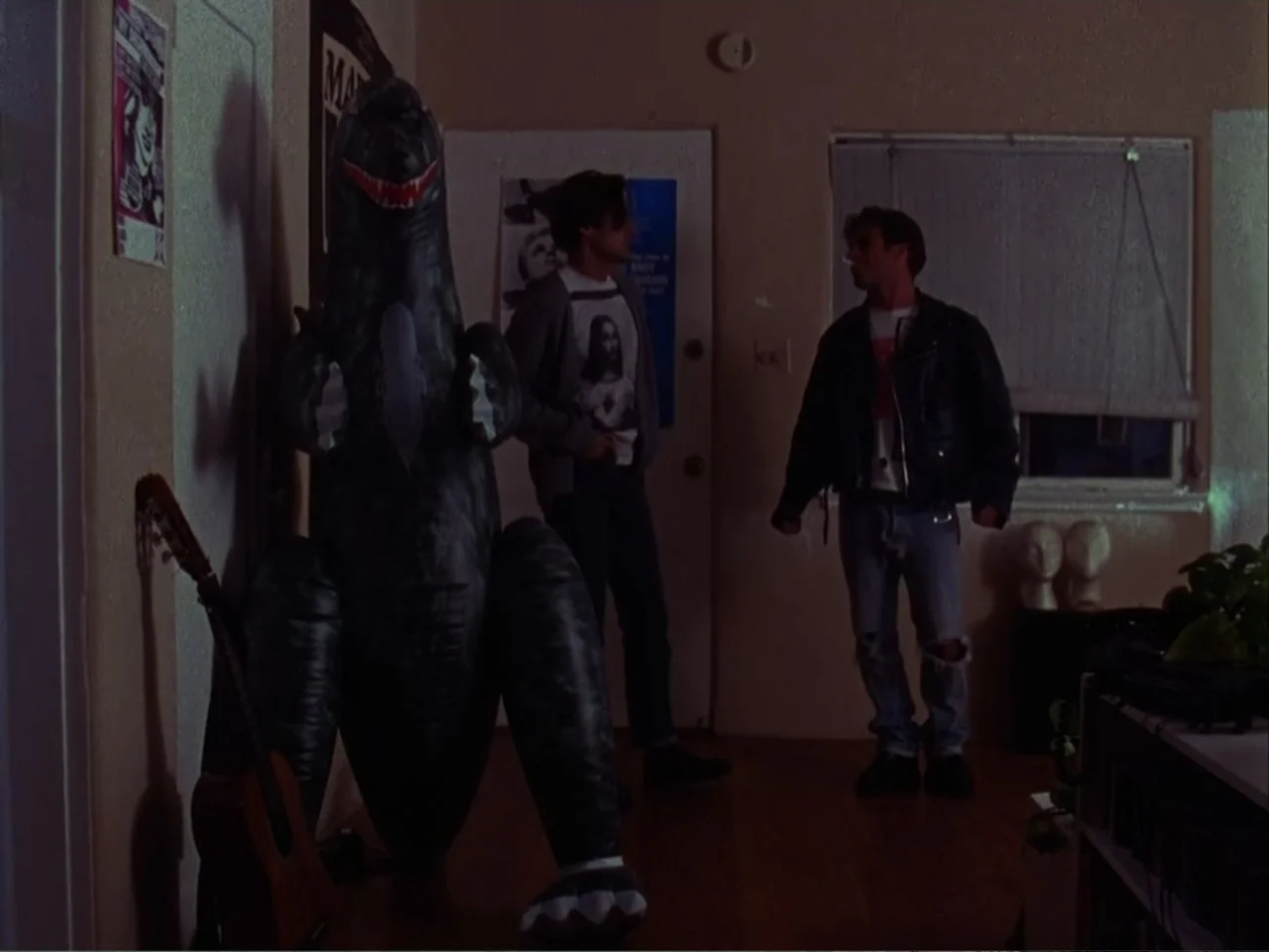 The Living End (1992) movie still: fullbody — A dimly lit room with plain walls adorned with posters and a guitar leaning against the wall. Two…