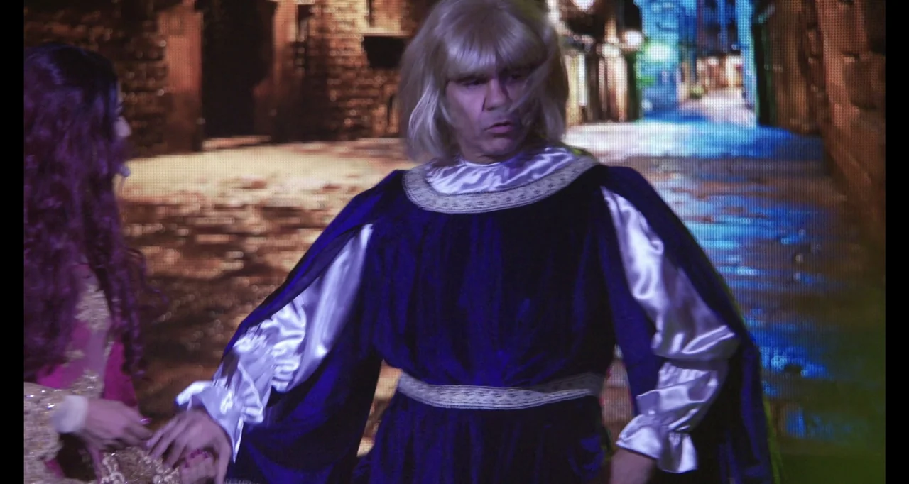 Paired Up (2019) movie still: medium — A man wearing a blonde wig and royal blue medieval tunic stands on a stage. A woman with purple hair stands…