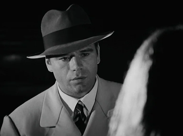 The American Soldier (1970) movie still: closeup — A man in a fedora and light suit stands in a dark urban environment. He observes an individual…