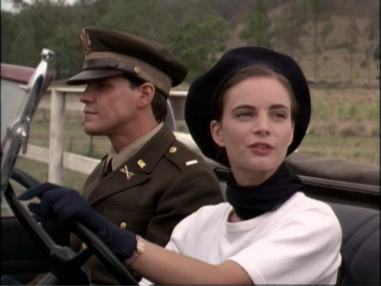 In Pursuit of Honor (1995) movie still: medium — A woman and a soldier sit in the front seats of an open-top vehicle. The background shows a rural…