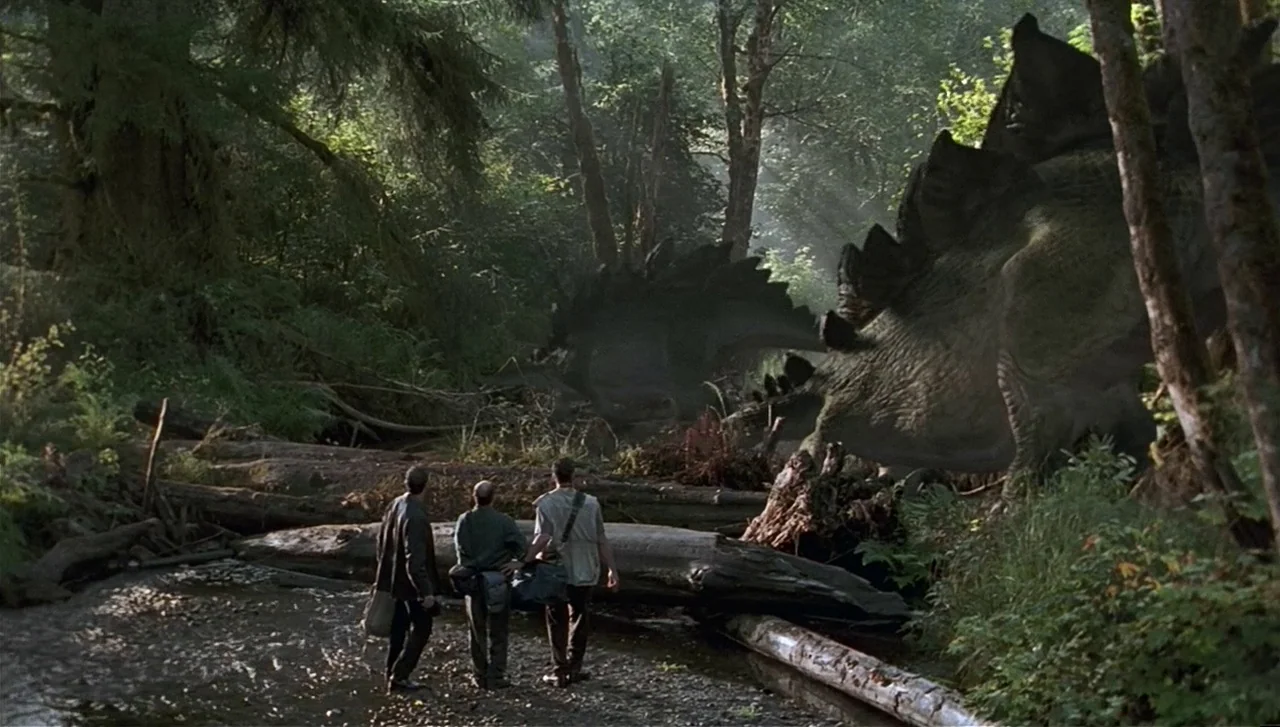 The Lost World: Jurassic Park (1997) movie still: wide — A lush woodland riverbank features fallen logs and dense ferns under tall trees. Three men…