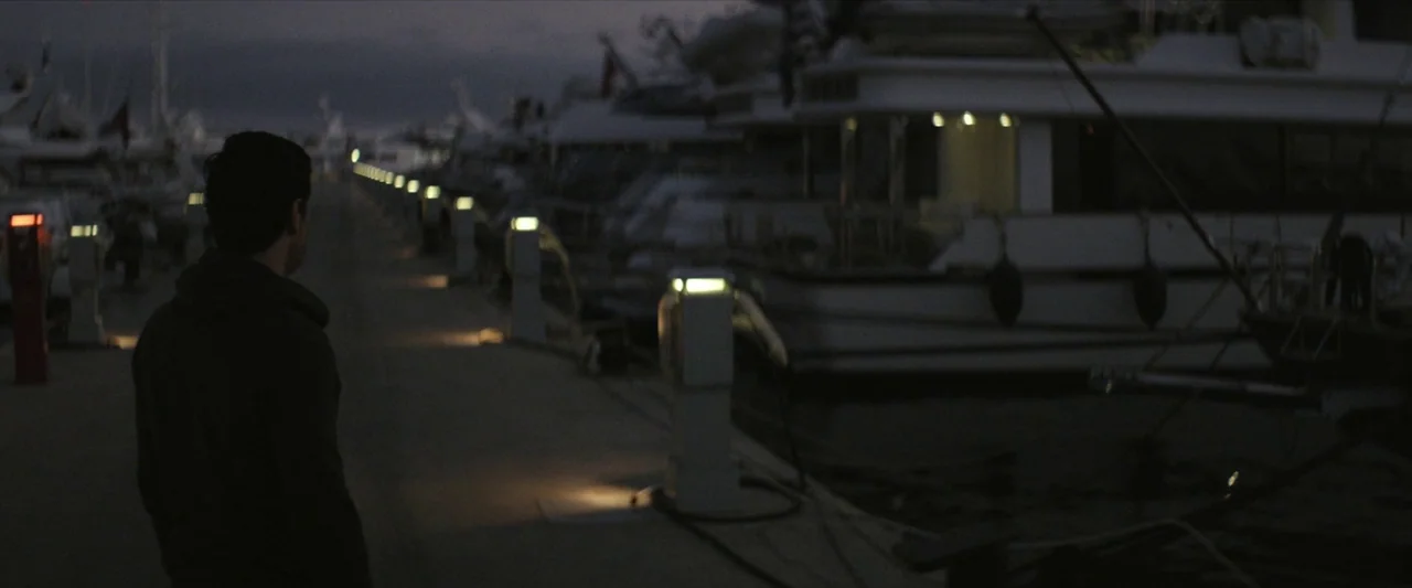 Chevalier (2015) movie still: medium — A dark, concrete walkway at a marina lined with lit utility pedestals at night. A lone man stands in the…