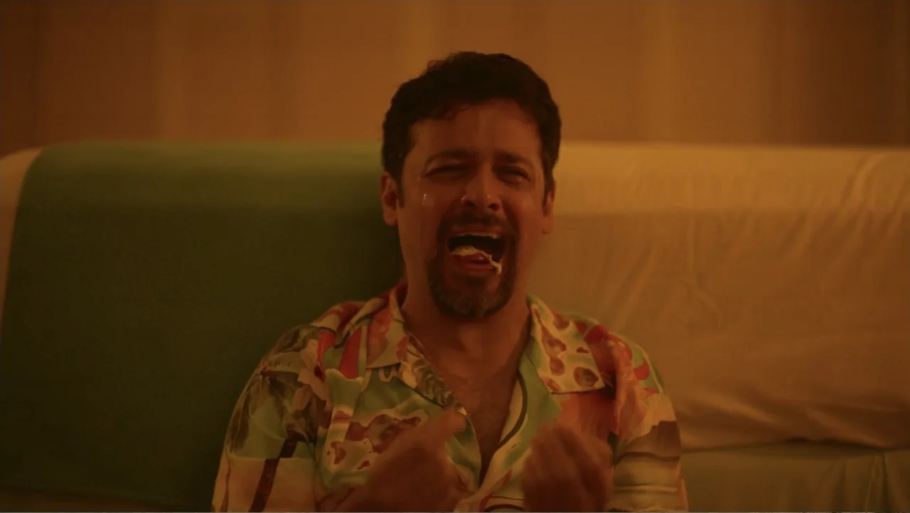 Double Pregnant 2 (2022) movie still: medium — A man sits on a couch in a dimly lit room with muted colors. The subject is crying with his mouth…